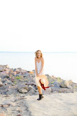 Caucasian blonde girl walking on rocky beach with hant in hands and wearing dress. Concept of summer vacations and fashion.の写真素材