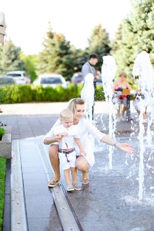 Young blonde mother sitting with little baby outside near fountain. Concept of walking in city and motherhood.の写真素材