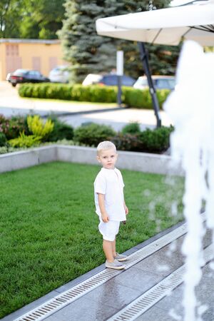 Little child standing on grass near fountain and wearing white clothes. Concept of childhood and summer.の写真素材