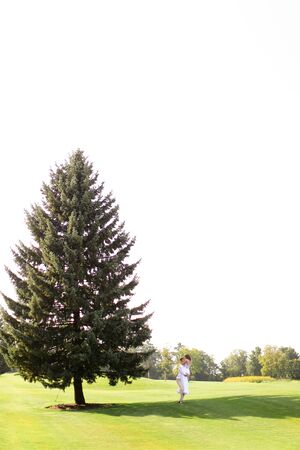 Young caucasian couple walking near green big spruce on grass in white sky background. Concept of evergreens and wedding.の写真素材