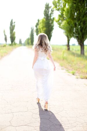 Back view of blonde caucasian bride wearing white dress walking on road. Concept of wedding photo session and fiancee.の写真素材
