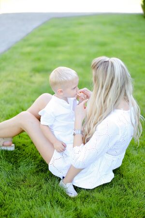 Young caucasian blonde mother sitting with little child on grass in yard. Concept of motherhood and summer vacations.の写真素材
