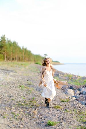 Caucasian blonde girl walking on shingle beach and wearing summer coat. Concept of seasonal fashion and summer vacations.の写真素材