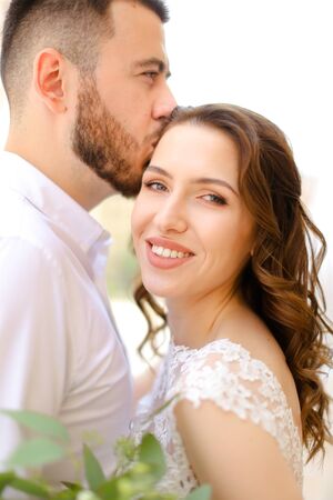 Happy caucasian groom kissing bride keeping bouquet of flowers. Concept of love and wedding photo session.の写真素材