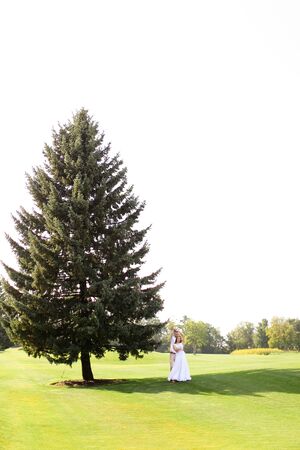 Young european couple walking near green big spruce on grass in white sky background. Concept of evergreens and wedding.の写真素材