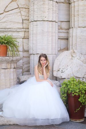 Caucasian pretty fiancee sitting near ancient columns and wearing white dress. Concept of bridal photo session and wedding.の写真素材
