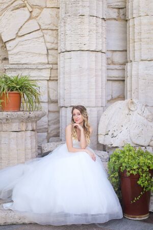 Caucasian nice fiancee sitting near ancient columns and wearing white dress. Concept of bridal photo session and wedding.の写真素材