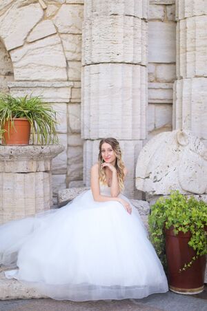 Caucasian beautiful fiancee sitting near ancient columns and wearing white dress. Concept of bridal photo session and wedding.の写真素材