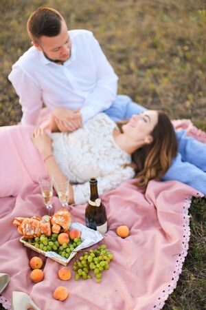 Young female person lying on man knees and pink plaid in steppe near fruits and bottle of red wine. oncept of picnic and relationship, romantic love.の写真素材