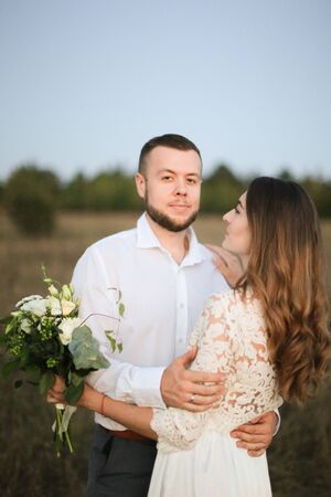 Close up handsome groom hugging bride with bouquet of flowers in field background. Concept of happy married couple and love.の写真素材