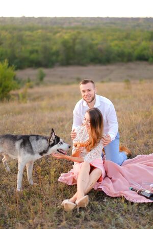Young european woman and man sitting on pink plaid near husky and resting outside. Concept of picnic on nature and dogs.の写真素材