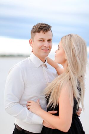 Young happy husband in white shirt hugging wife wearing black dress in winter steppe. Concept of winter photo session, relationship and love, happy couple.の写真素材
