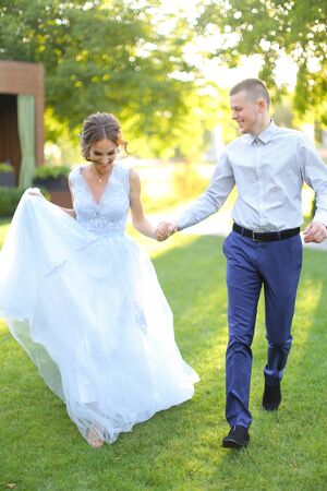 Cacuasian fiancee walking with groom in park and holding hands, wearing white dress. Concept of wedding and bridal photo session in open air.の写真素材