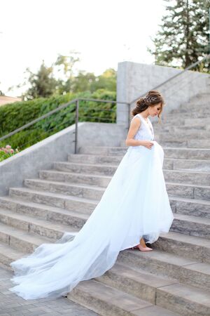 Back view of caucasian young bride walking on concrete stairs and wearing white dress. Concept of bridal fashion and wedding vogue.の写真素材