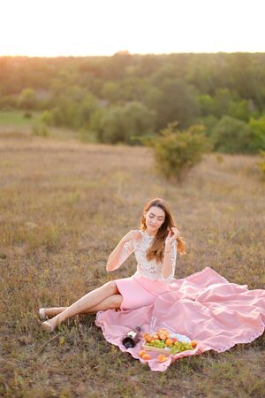 Young girl sitting on pink plaid near bottle of red wine and fruits. Concept of resting in open air and summer picnic.の写真素材