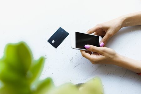 Female hands keeping credit card and smartphone in white and green leaves background with copyspace. Concept of shopping online.の写真素材