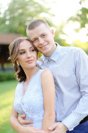 Happy caucasian groom hugging fiancee in park, sun rays. Concept of bridal couple and wedding photo session in open air.の写真素材