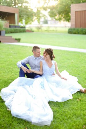 Caucasian bride with handsome groom sitting on grass in park, wearing white amazing dress. Concept of bridal photo session in open air, beautiful fiancee.の写真素材