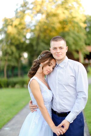 Young cute fiancee standing and hugging with caucasian groom in park. Concept of happy couple and wedding photo session outside.の写真素材