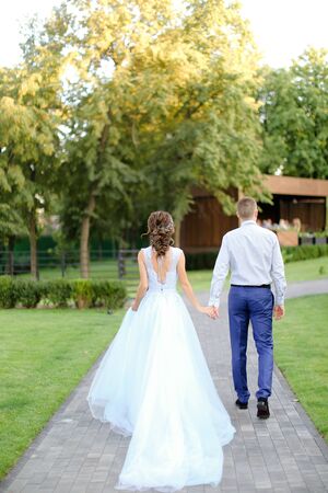 Back view of caucasian fiancee and groom going outdoors. Concept of wedding photo session outside and happy bridal couple walking in garden.の写真素材