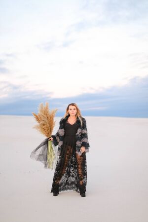 Lady wearing fur coat and transparent black dress standing in snowed steppe with dried plants bouquet. Concept of fashion and female person, winter photo session.の写真素材