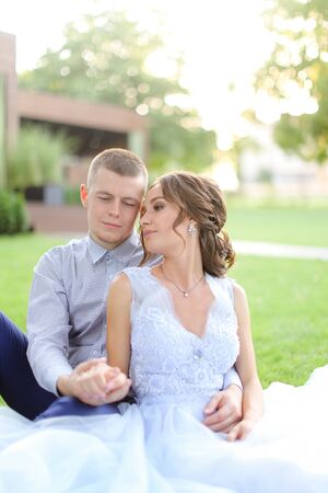 Happy groom hugging bride and sitting on grass in park. Concept of married couple and bridal photo session in open air.の写真素材