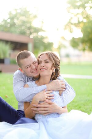 Happy groom hugging nice bride and sitting on grass in park. Concept of married couple and bridal photo session in open air.の写真素材