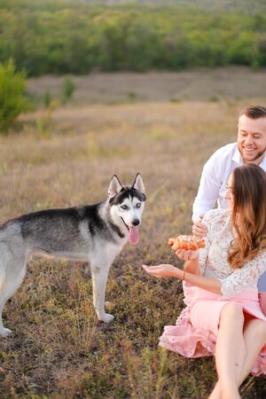 Young woman and man sitting on grass plaid near husky and resting outside. Concept of picnic on nature and dogs.の写真素材
