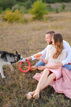 Young happy woman and man sitting on grass plaid and playing with husky. Concept of picnic on nature and dogs.の写真素材