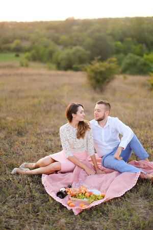 Young european wife and husband sitting on pink plaid in field. Concept of picnic in open air and relationship, love.の写真素材