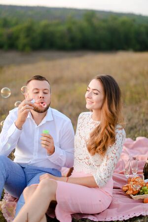 Young boy blowing soap bubbles near european girl and sitting on pink plaid in steppe. Concept of happy couple, romantic love and picnic on nature.の写真素材
