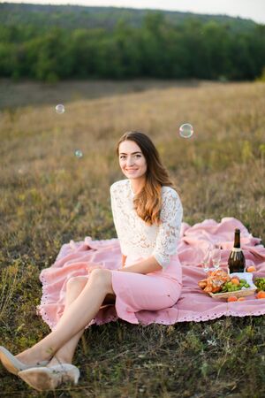Young happy woman sitting on plaid with bottle of wine and fruits, soap bubbles. Concept of beauty nad picnic on nature.の写真素材