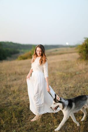 Caucasian woman walking in steppe with dog husky and wearing white dress. Concept of nature, bridal photo session on nature and beauty, pets.の写真素材