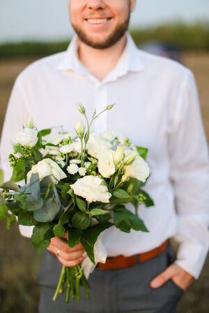 Young caucasian groom standing with bouquet of white flowers in steppe, wearing shirt. Concept of wedding photo session and romantic male person.の写真素材