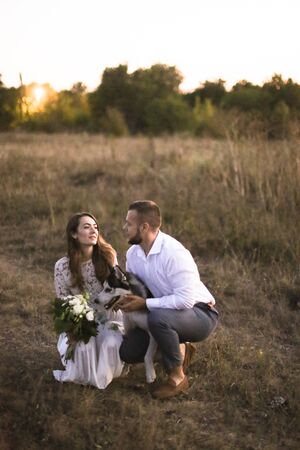 Bride and happy groom sitting on grass with dog in field. Concept of wedding photo session on nature and pets.の写真素材