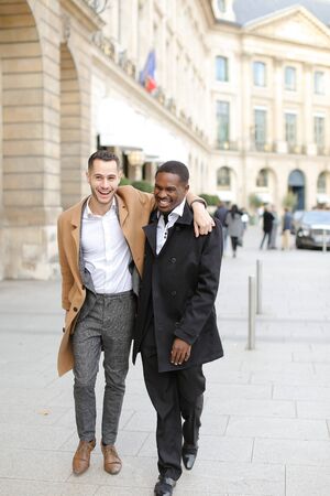 Caucasian smiling man in suit walking with afroamerican male person and hugging in city. Concept of happy gays and strolling.の写真素材