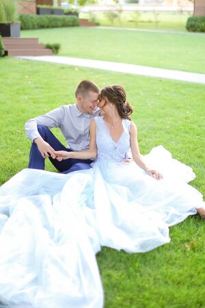 Caucasian bride with groom sitting and kissing on grass in park, wearing white amazing dress. Concept of bridal photo session in open air, beautiful fiancee.の写真素材