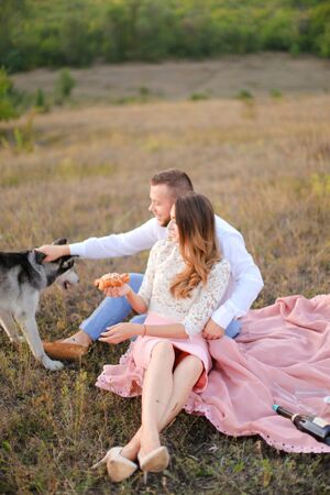 Young happy girl and man sitting on grass plaid and playing with husky. Concept of picnic on nature and dogs.の写真素材