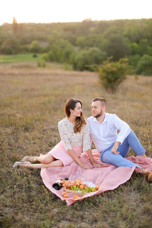 Young american wife and husband sitting on pink plaid in field. Concept of picnic in open air and relationship, love.の写真素材