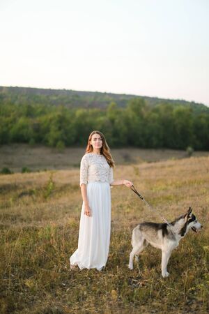 Caucasian pretty woman walking in steppe with dog husky and wearing white dress. Concept of nature, bridal photo session on nature and beauty, pets.の写真素材