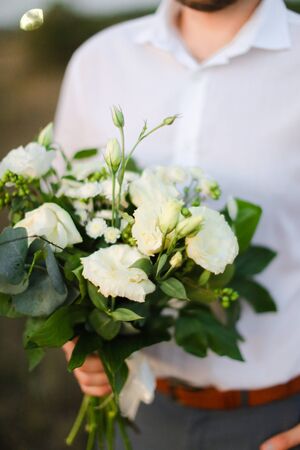 Young caucasian man standing with bouquet of white flowers in steppe, wearing shirt. Concept of wedding photo session and romantic male person.の写真素材
