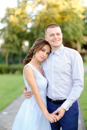 Young beautiful fiancee standing and hugging with caucasian groom in park. Concept of happy couple and wedding photo session outside.の写真素材