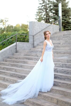 Caucasian pretty bride standing on concrete stairs and wearing white dress. Concept of bridal fashion and wedding vogue.の写真素材