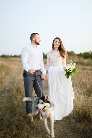 Groom walking with beautiful fiancee and dog in steppe, holding hands. Concept of happy married couple and love.の写真素材