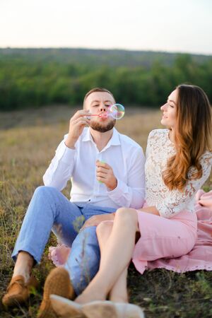 Young guy blowing soap bubbles near caucasian girl and sitting on pink plaid in steppe. Concept of happy couple, romantic love and picnic on nature.の写真素材