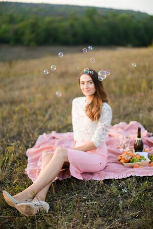 Young happy woman sitting on plaid with bottle of wine and fruits, focus on soap bubbles. Concept of beauty nad picnic on nature.の写真素材