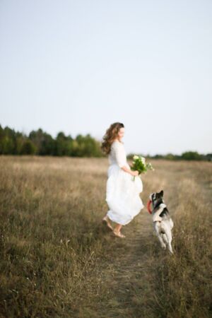 Fiancee in white dress walking in field with dog and bouquet of flowers. Concept of bridal photo session with pet dogs and nature.の写真素材