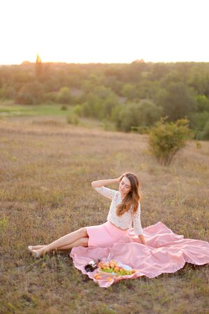 Young caucasian girl sitting on pink plaid near bottle of red wine and fruits. Concept of resting in open air and summer picnic.の写真素材