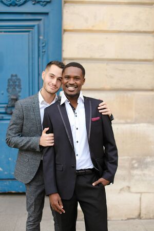 Two boys, caucasian and afro american, wearing suits standing near building and hugging. Concept of gays and lgbt.の写真素材