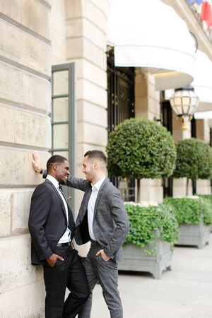 Afro american and european gays standing near building and wearing suits and talking outside. Concept of lgbt and walking in city.の写真素材